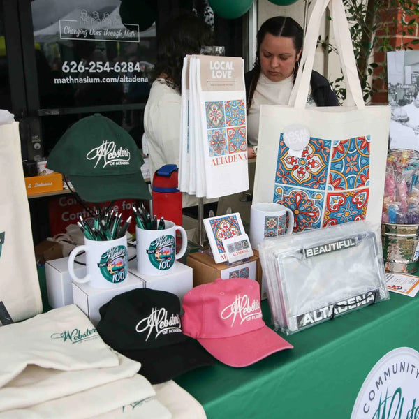 Webster's Pharmacy 100th anniversary tote bags, caps and mugs displayed on a green booth table