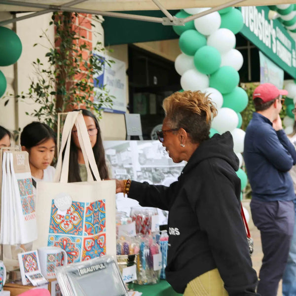 Shoppers browsing tote bags under the balloon arch outside Webster's Pharmacy